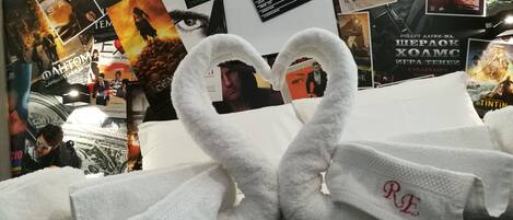 A room with a bed covered by a white blanket, a wall with movie posters, and a white towel with a heart-shaped design.