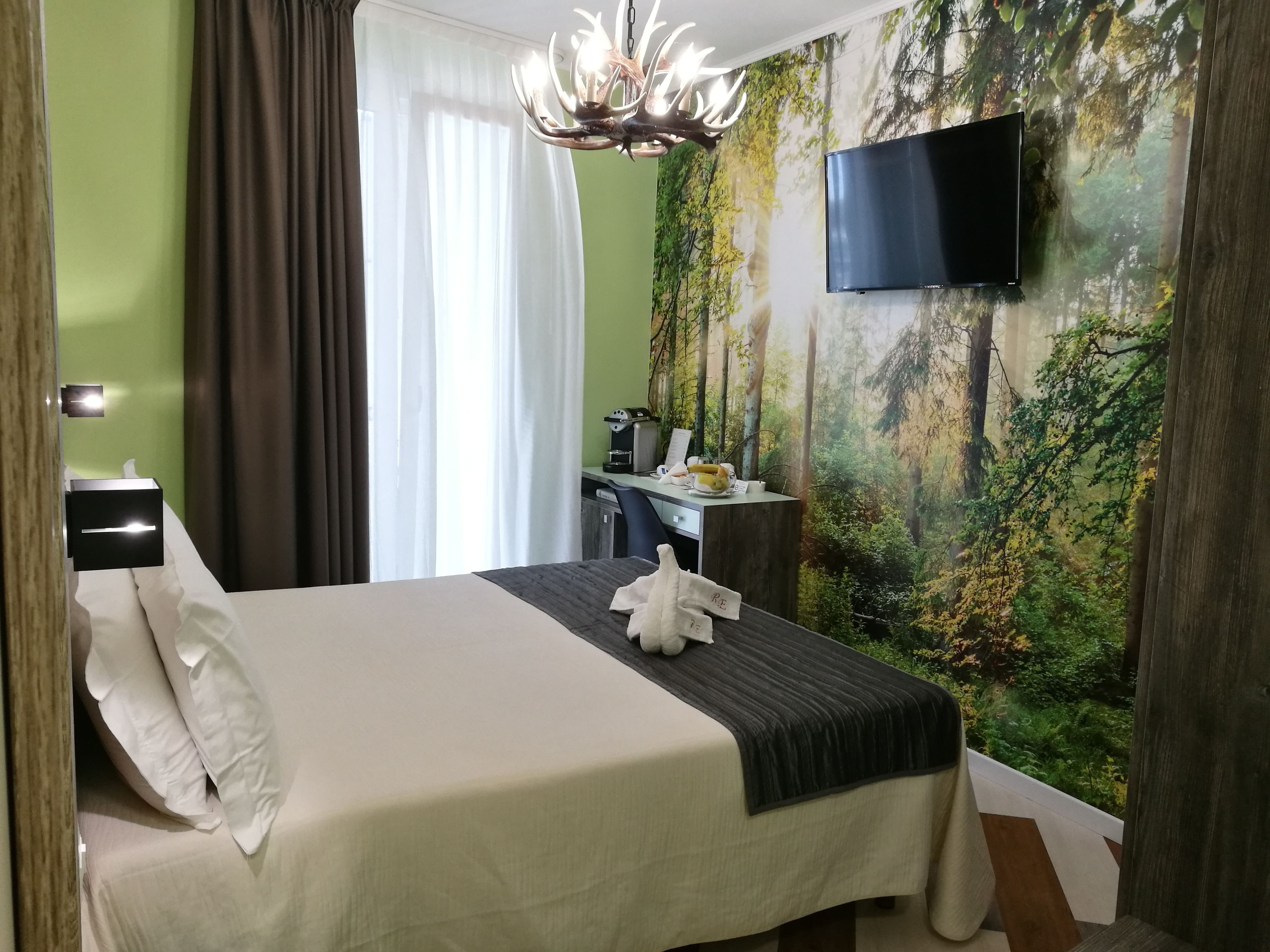 A hotel room with a bed, a desk, a chair, a TV, and a forest mural on the wall.