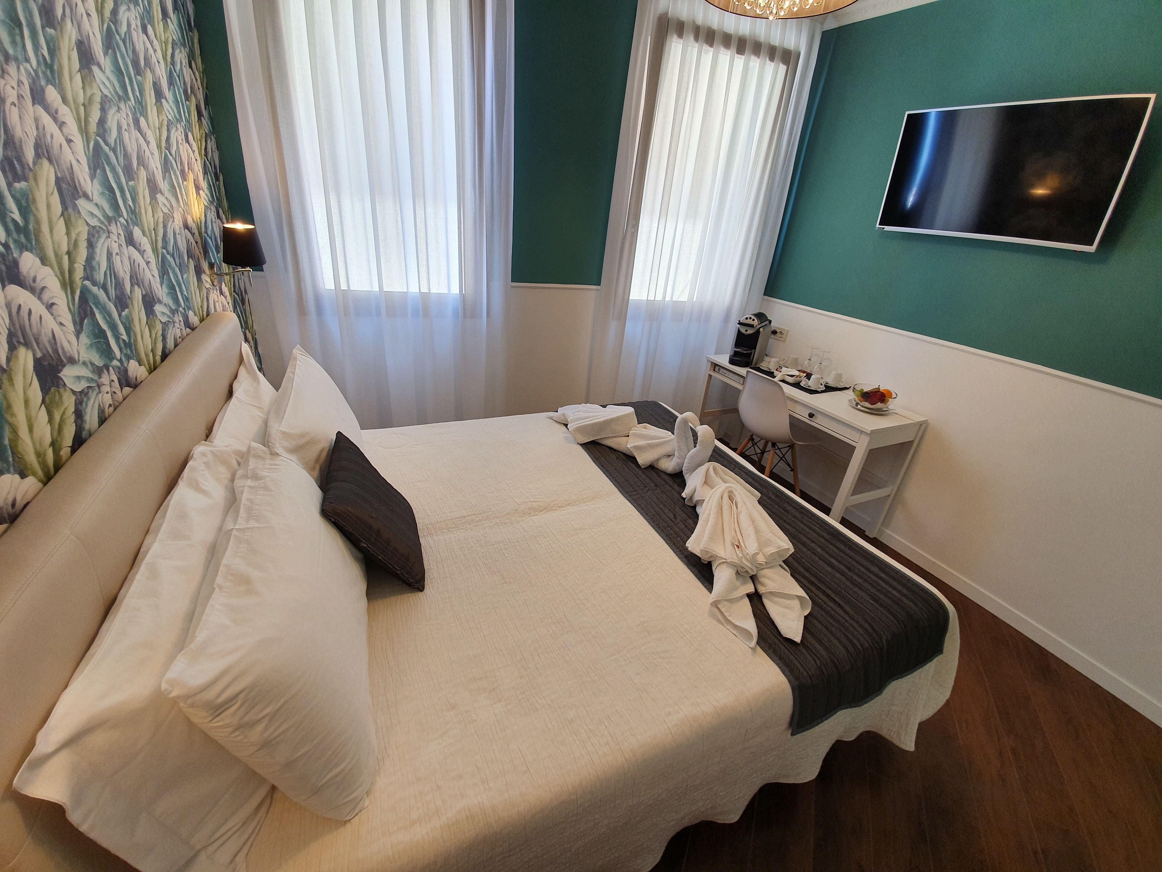 A neatly made bed with towels, a desk with a coffee maker, and a TV on the wall.
