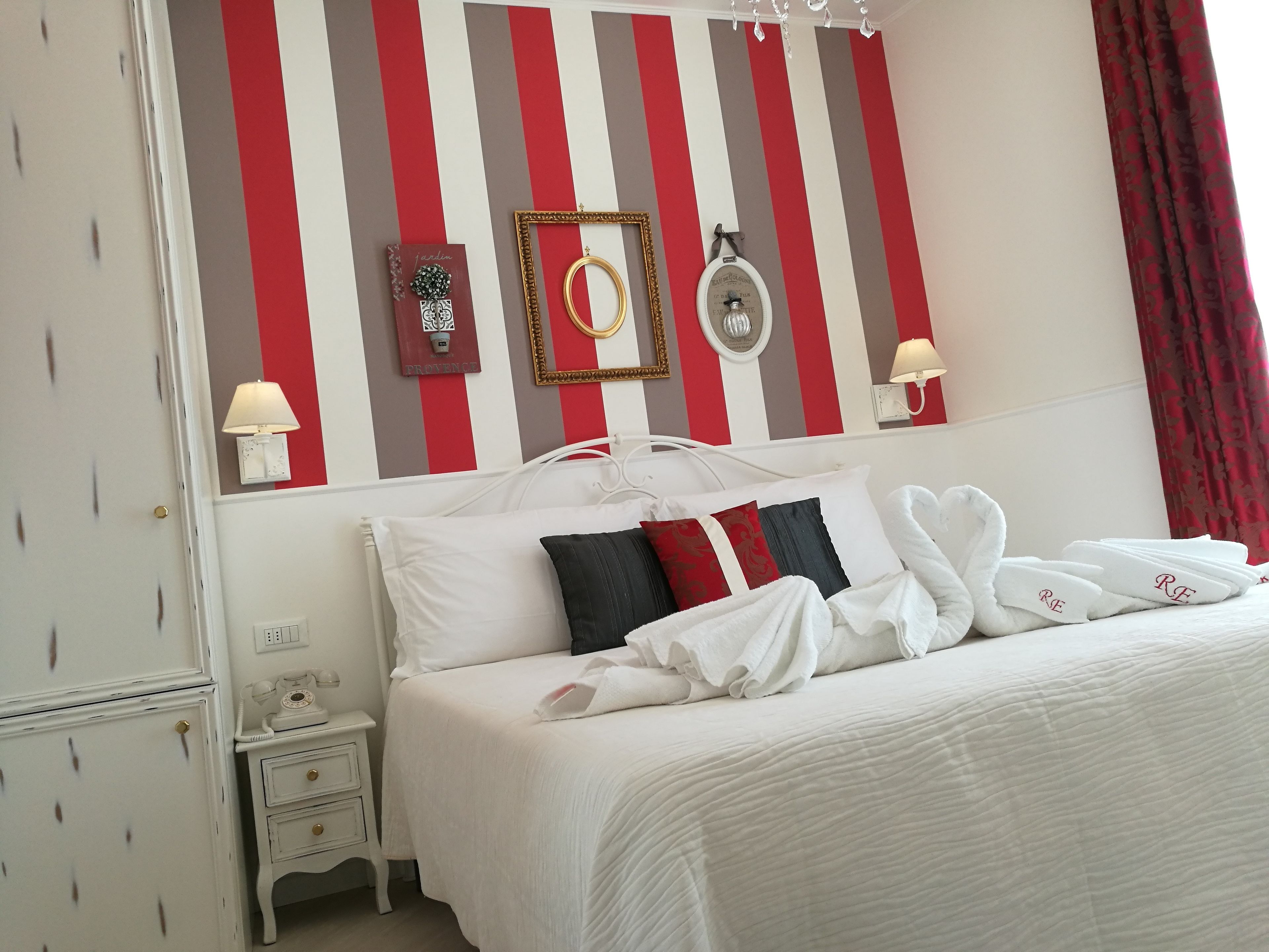Ein ordentlich bezogenes Bett mit weißen Leinen und Handtüchern, ein Nachttisch mit Lampe sowie eine Wand mit gestreifter Tapete und gerahmten Dekorationen.