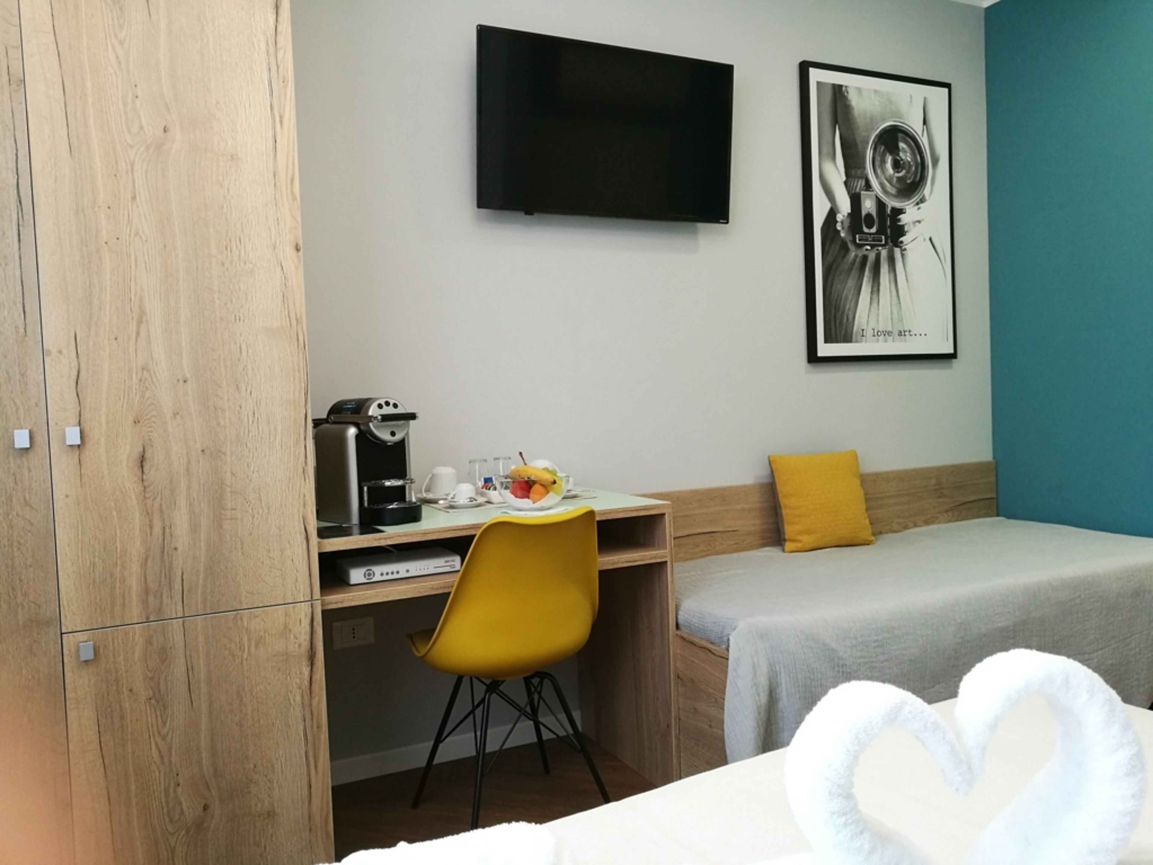 Een hotelkamer met een bed, een bureau met stoel, een koffieapparaat, een flatscreen televisie en een lijst met een kunstwerk aan de muur.