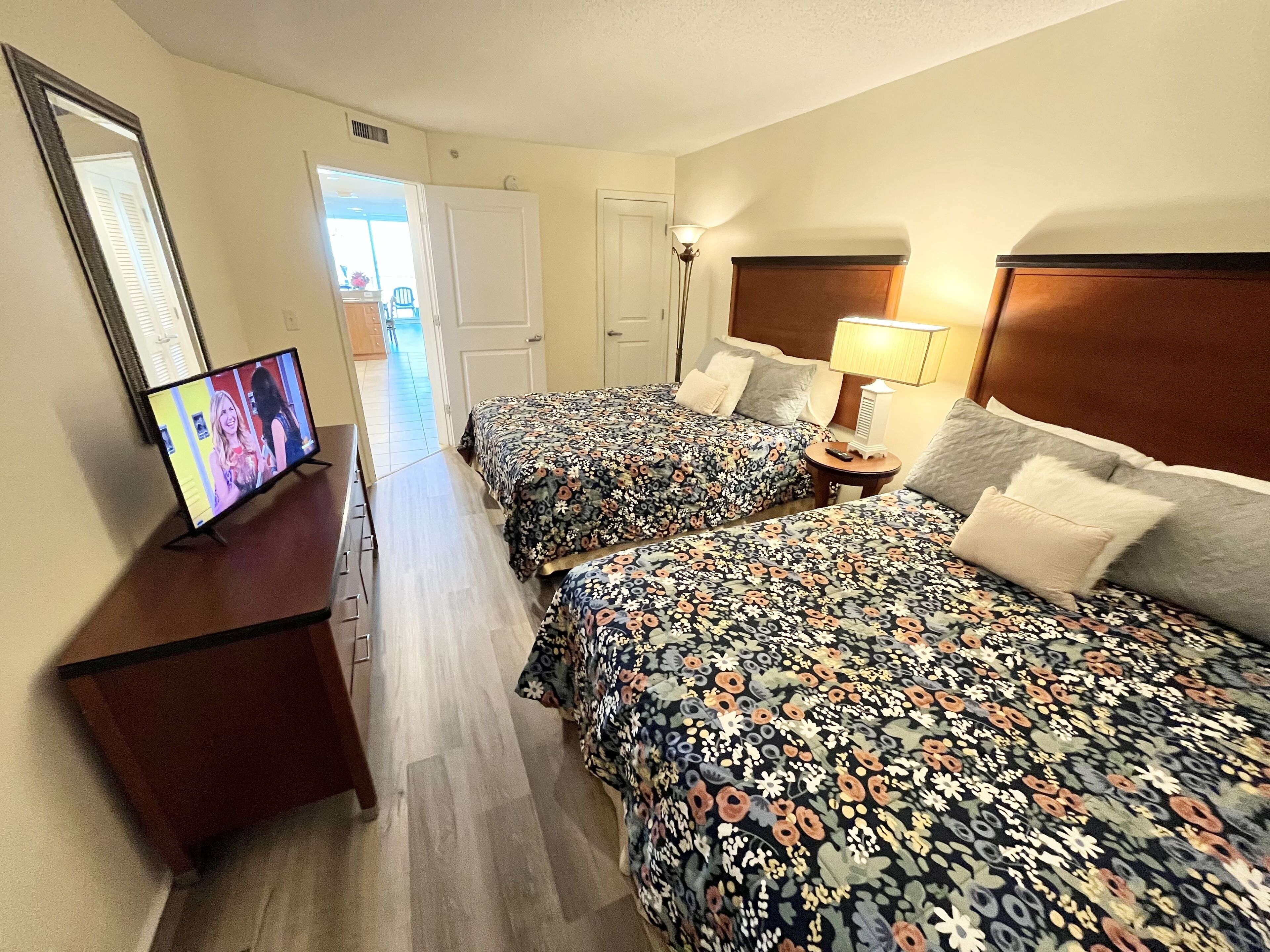 A hotel room with two beds, a TV on a stand, a bedside table with a lamp, and a door leading to another room.