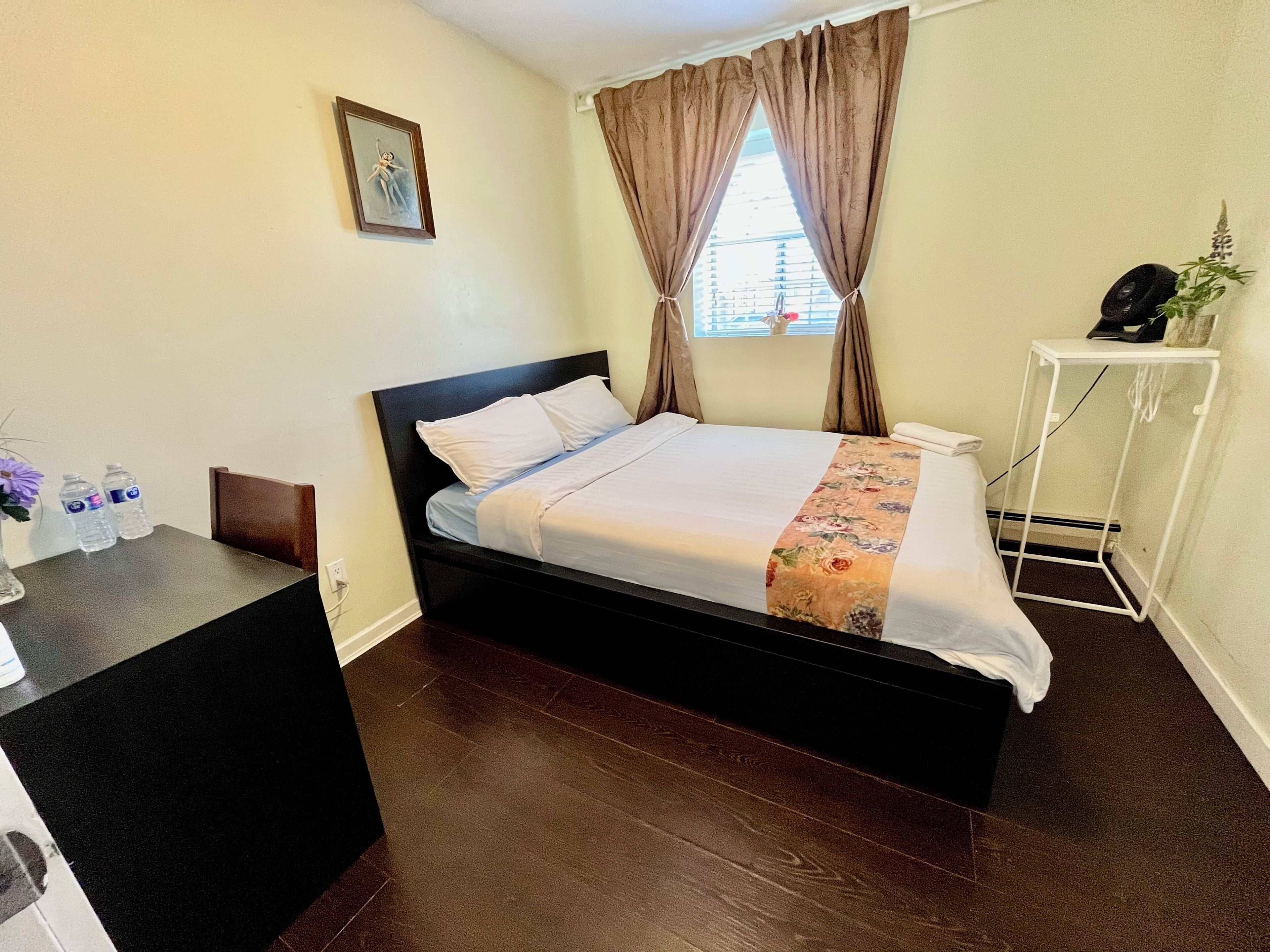 A hotel room with a bed, a desk, a chair, a small table, a window with curtains, and a wall-mounted picture.