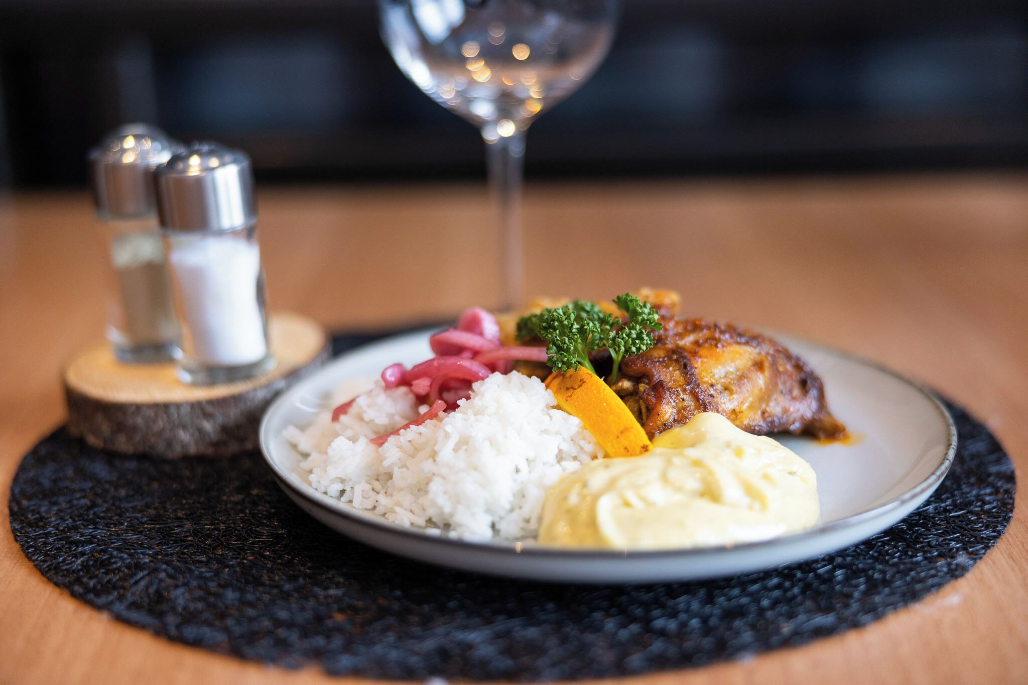 A plate of food with rice, a meat dish, and a side of sauce, accompanied by a glass of wine and salt and pepper shakers on a wooden table.