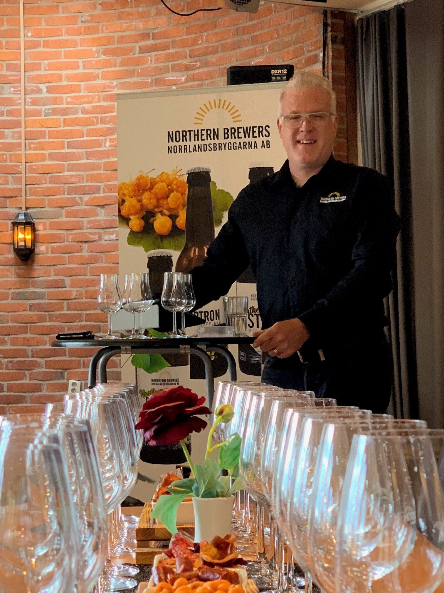 A man standing behind a table with glasses and bottles, a brick wall, and a sign for Northern Brewers.