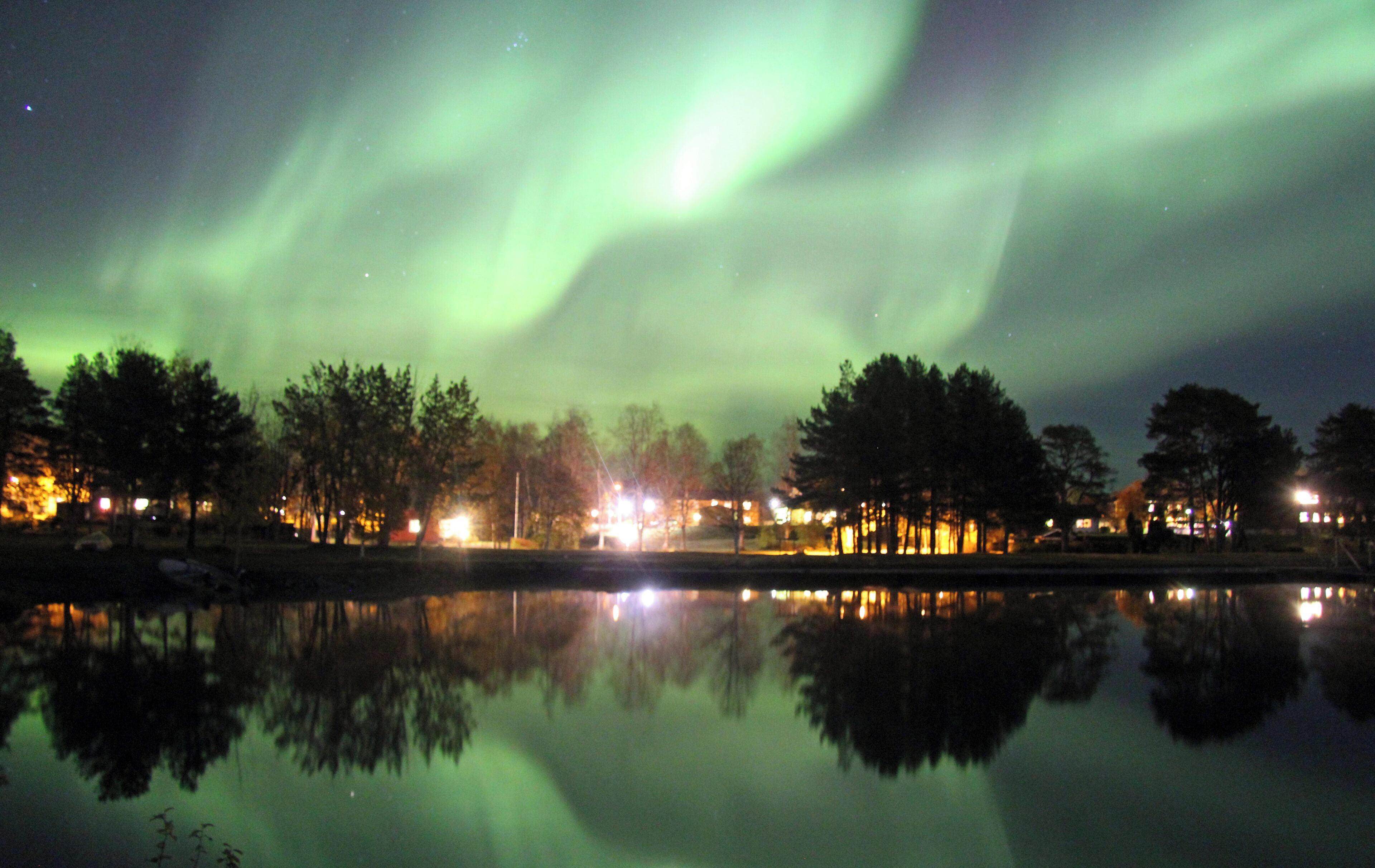 A nighttime view of the Northern Lights over a calm lake with reflections of trees and lights from a nearby settlement.