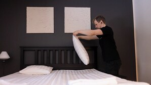 Uma pessoa ajustando um travesseiro em uma cama em quarto de hotel, com uma cabeceira escura e duas obras de arte simples nas paredes.