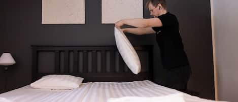 A person adjusting a pillow on a bed in a hotel room with a dark headboard and two plain wall art pieces.