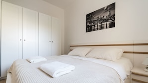 A neatly made bed with a white blanket and a folded towel, a bedside table with a lamp, and a cityscape painting on the wall. - Yafo 35 - Isrentals (Jerusalem)