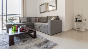 A modern living room with a grey sectional sofa, a coffee table with snacks, a large window, and a white ladder against the wall. - Yafo 35 - Isrentals (Jerusalem)