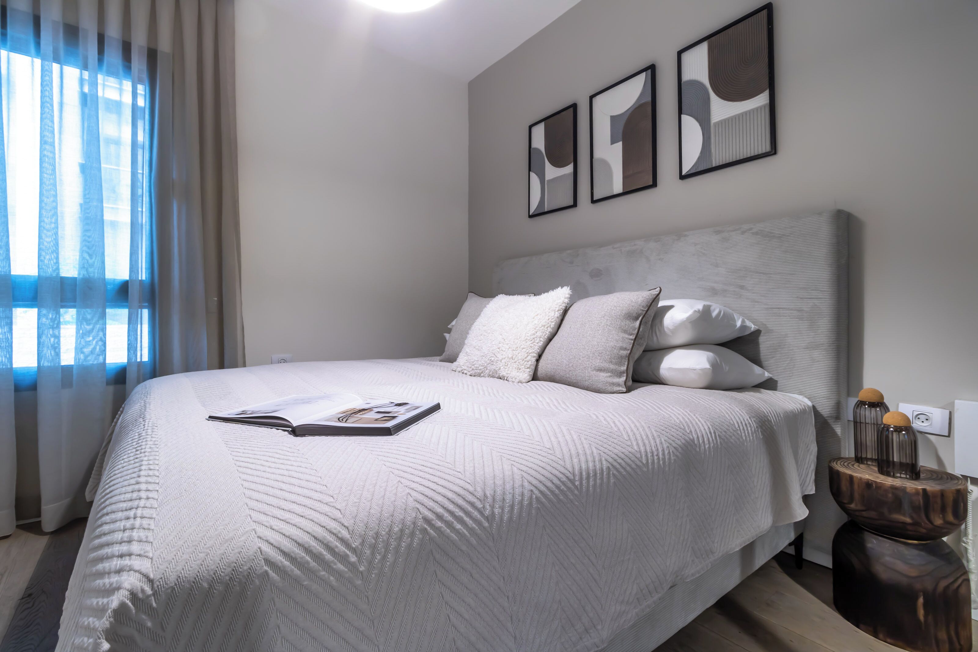A modern bedroom with a large bed, a bedside table, and a window with sheer curtains.