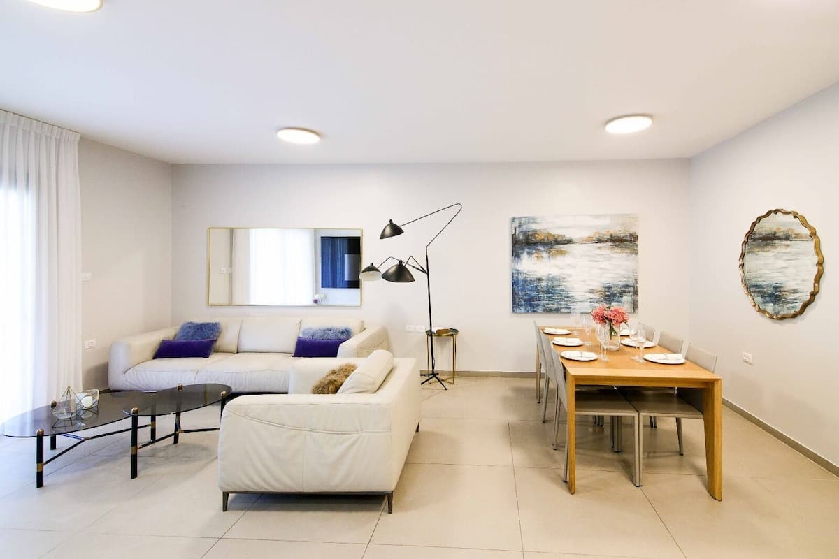 Un salon moderne avec un canapé blanc, une table basse en verre, une table à manger pour quatre personnes et un grand miroir au mur.