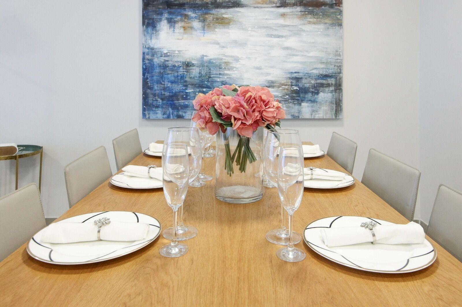 A dining table set for six with white plates, clear wine glasses, and a centerpiece of pink flowers.