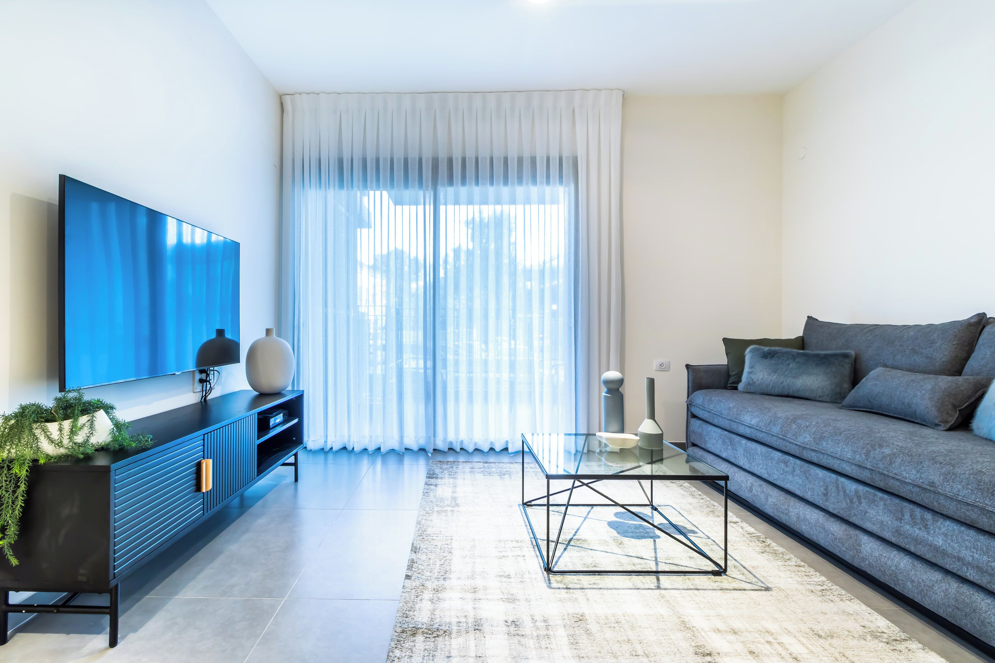 Una sala de estar moderna con televisor de pantalla plana, un sofá, una mesa de centro de vidrio y un amplio ventanal con cortinas translúcidas.