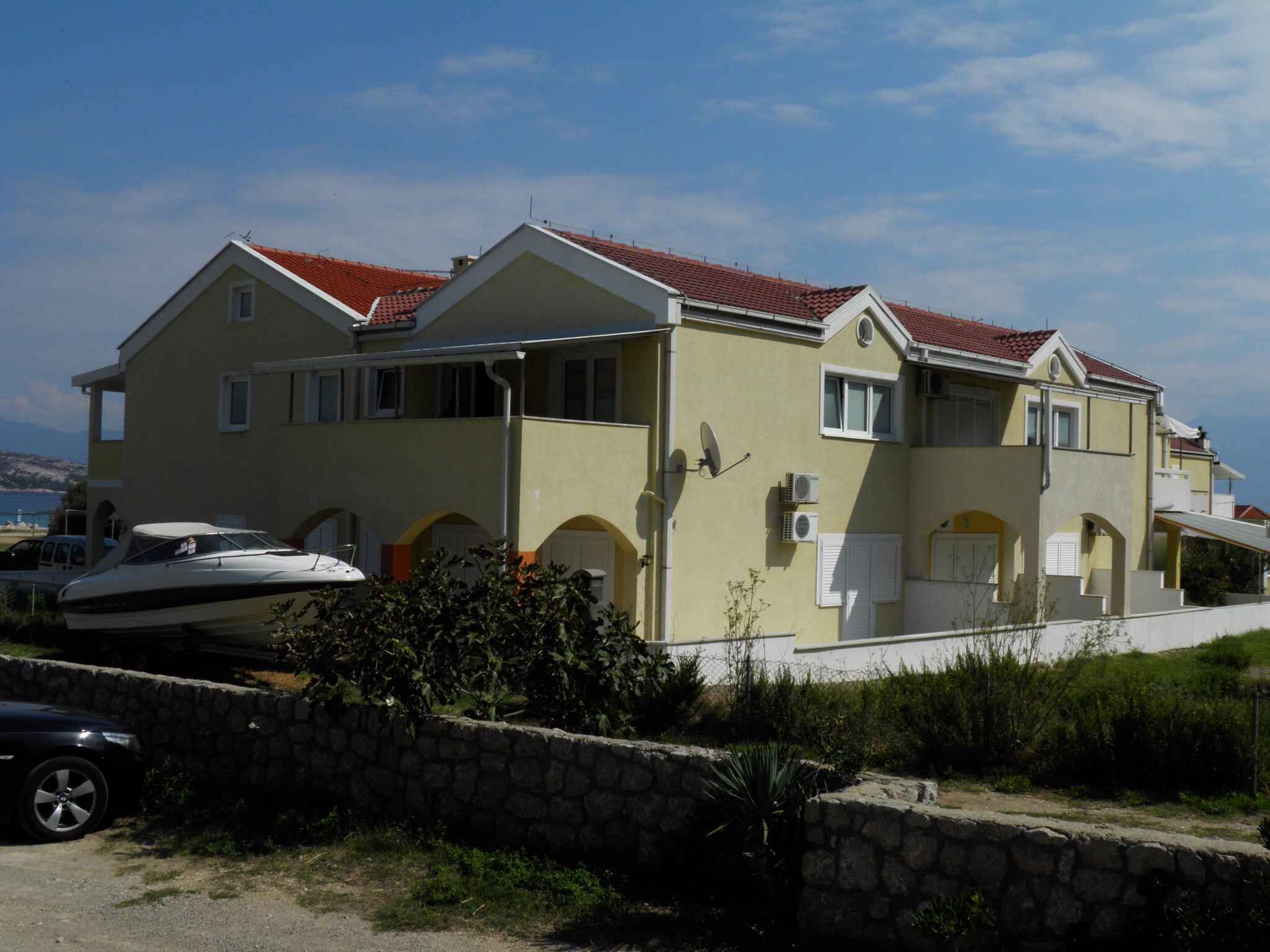 A two-story house with a red-tiled roof, a stone wall, and a boat parked in front.