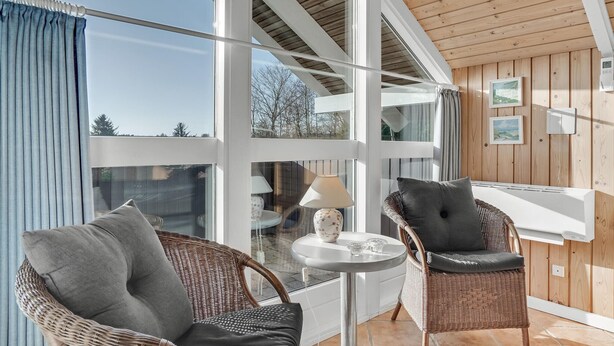 Et moderne værelse med fletstole, et lille bord med lampe, og store vinduer med udsigt til det fri.