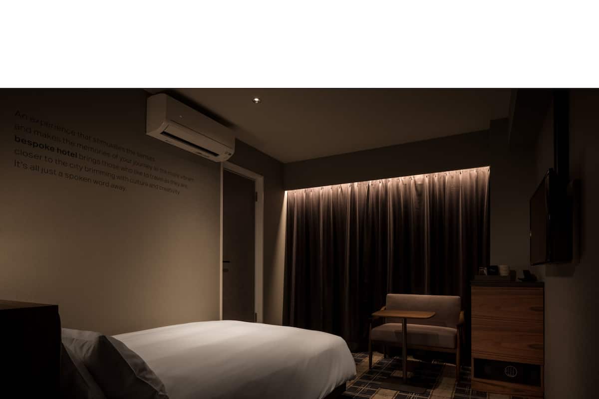 Une chambre d’hôtel faiblement éclairée, avec un lit, une chaise, une petite table et un climatiseur fixé au mur.