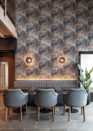 Interior de restaurante con diseño moderno, que presenta una pared con textura, dos lámparas colgantes y una zona de estar con asientos tipo banca tapizados en gris y sillas de madera.