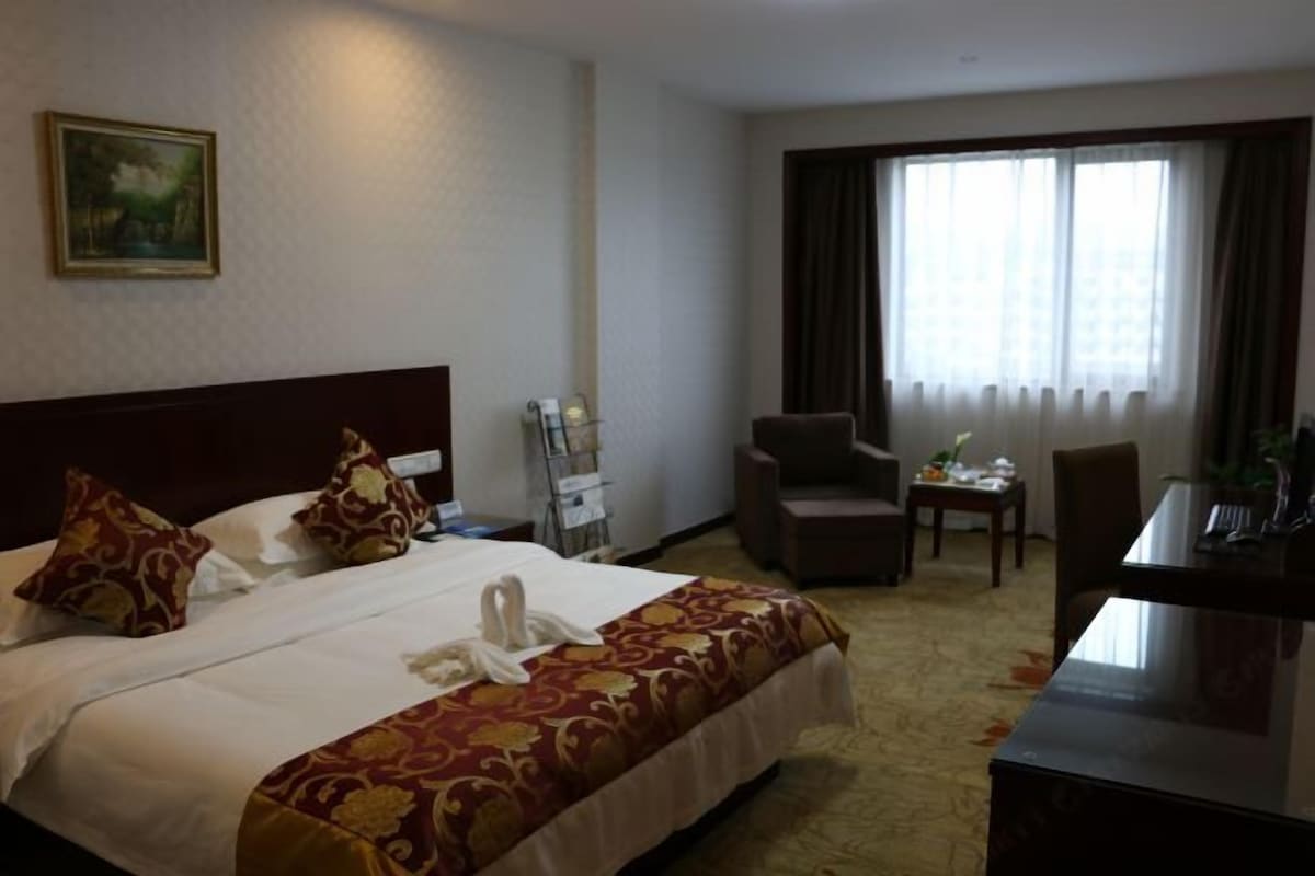 A hotel room with a bed, a desk, a chair, a window with curtains, and a painting on the wall.
