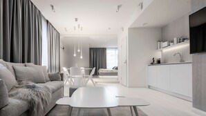 Een moderne woonkamer met een bank, een salontafel en een eethoek met stoelen. Er is een keuken met een spoelbak en een televisie aan de muur bevestigd.