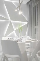 Un comedor moderno con una mesa blanca para dos, diseño geométrico en la pared y un jarrón con plantas.