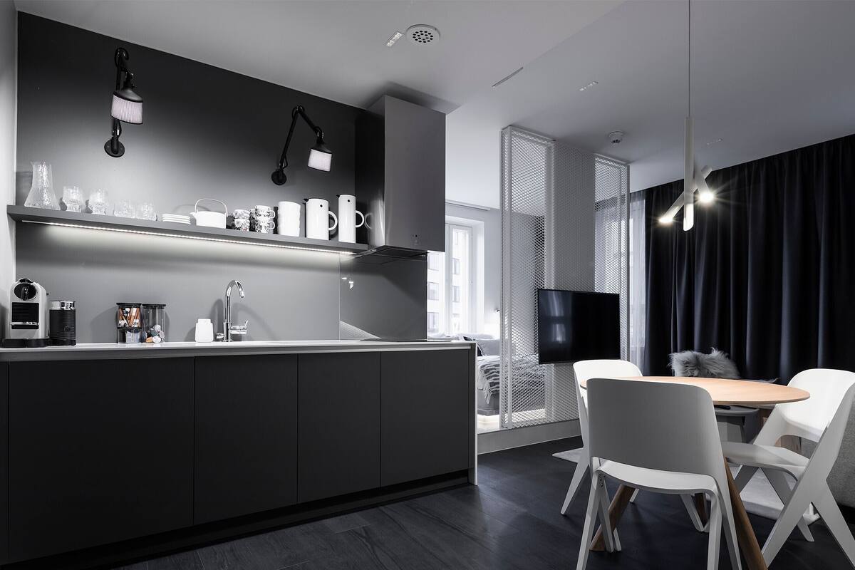 Une cuisine moderne avec un plan de travail foncé, une machine à café et un évier. On y trouve également un coin repas avec une table et des chaises, ainsi qu’un téléviseur fixé au mur.