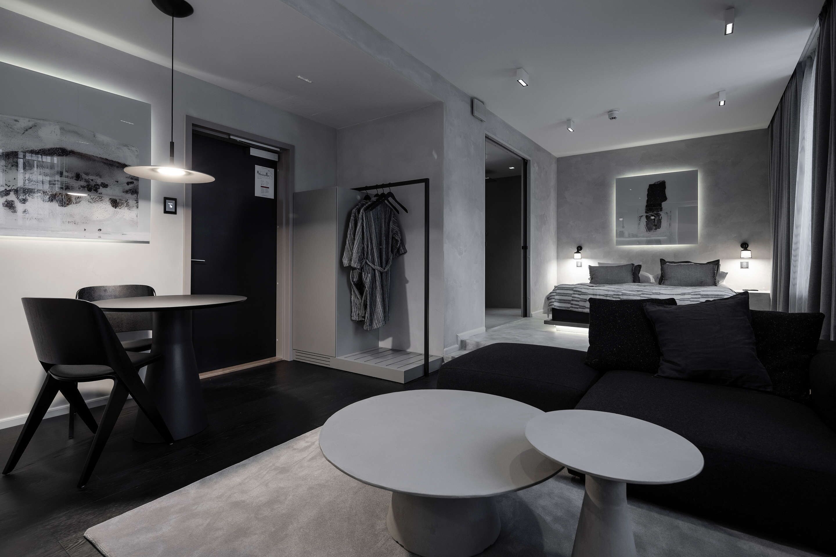 Habitació d'hotel moderna amb un sofà de color fosc, una taula rodona de dinar, una taula petita blanca i una bata penjada.