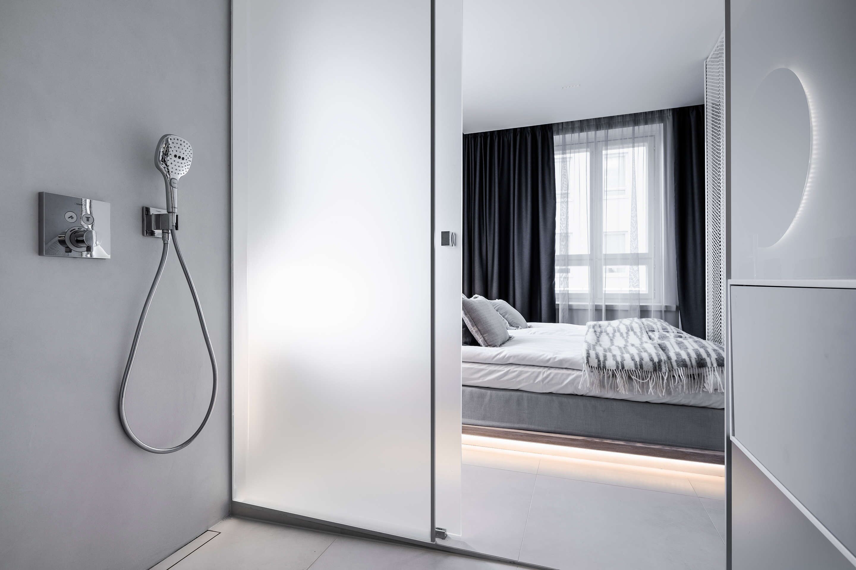 Un baño moderno con ducha, inodoro y bidé. La habitación tiene un ventanal grande con cortinas y una cama con cubrecama blanco y gris.