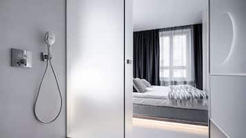Un baño moderno con ducha, inodoro y bidé. La habitación tiene un ventanal grande con cortinas y una cama con cubrecama blanco y gris.