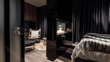 A modern hotel room with a large bed, a seating area with a chair and a small table, and a fireplace.