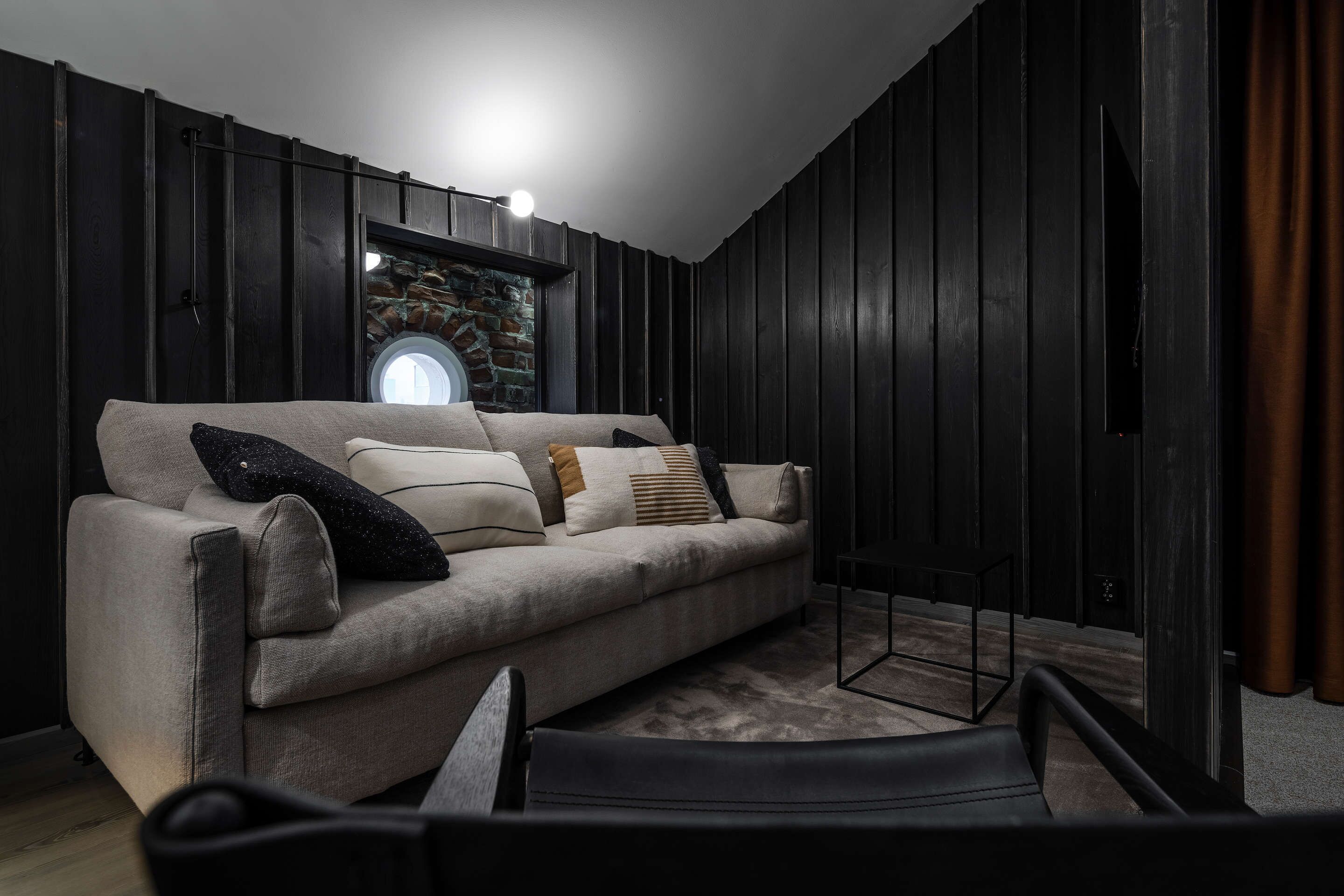 A modern living room with a beige sofa, black and white cushions, a round window, and dark wooden walls.