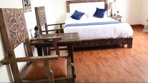A wooden bed with white bedding and blue pillows, a wooden nightstand with a lamp, and a wooden chair with a cushion.