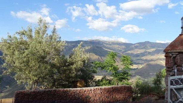 A stone wall with a metal gate, a rustic building with a chimney, and a mountainous landscape with trees.