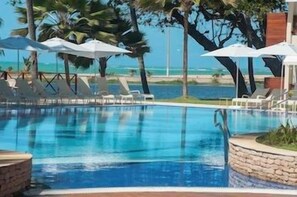 A swimming pool with lounge chairs and umbrellas, surrounded by palm trees and a view of the ocean. - Jatiuca Resort (MACEIO)