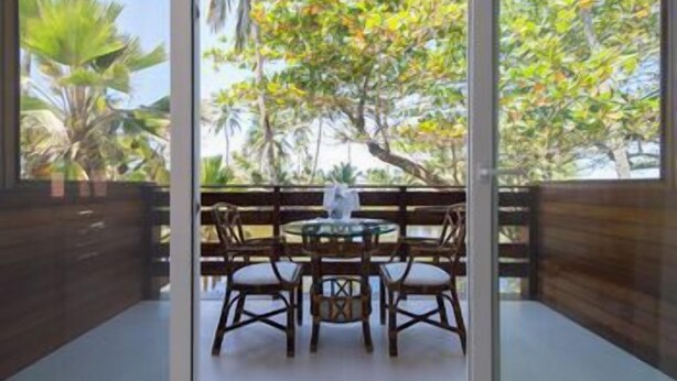 A room with a glass door leading to a balcony with a table and chairs, surrounded by tropical vegetation.