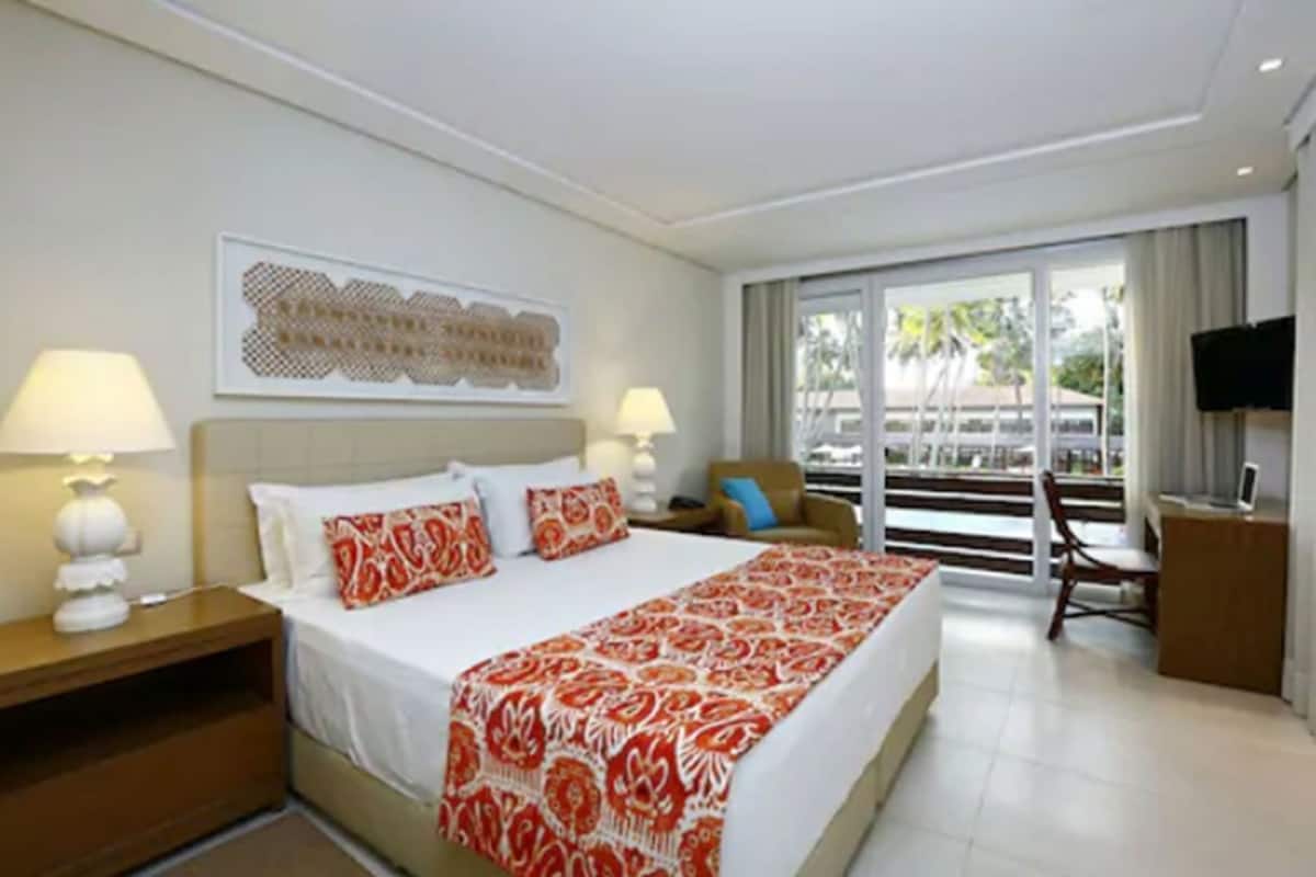 A hotel room with a bed, two bedside lamps, a television, a chair, and a view of the outdoors.