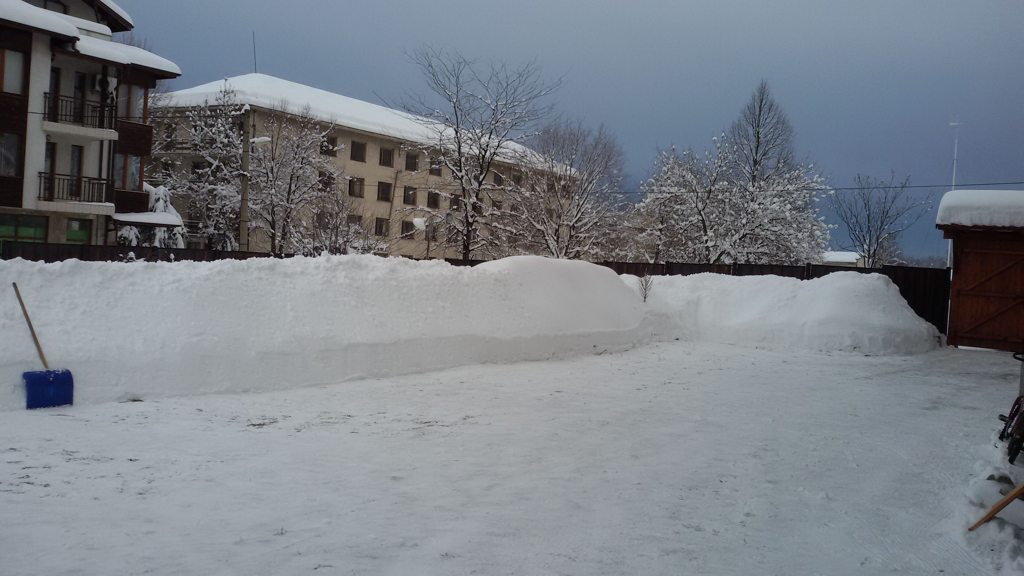 Ein verschneiter Innenhof mit einem großen Schneehaufen, einer blauen Schaufel und einem Gebäude mit Balkonen im Hintergrund.
