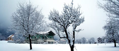 A snow-covered landscape with a large building featuring multiple floors and a balcony, surrounded by trees.