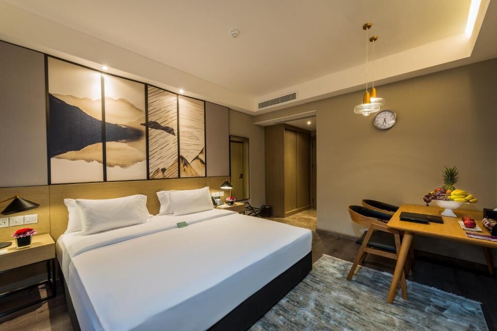 A hotel room with a large bed, a desk with a chair, a clock on the wall, and a dining table with fruits.