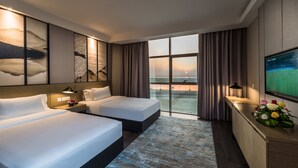 A hotel room with two beds, a large TV, a desk with a vase of flowers, and a view of the sunset through the window. - Boudl Al Corniche (Dammam)