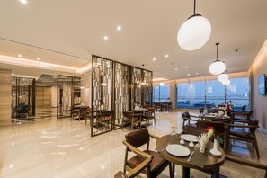 A modern dining area with a high ceiling, recessed lighting, and a view of the cityscape through large windows. - Boudl Al Corniche (Dammam)