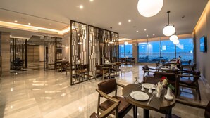 A modern dining area with a high ceiling, contemporary furniture, and large windows offering a view of the cityscape. - Boudl Al Corniche (Dammam)