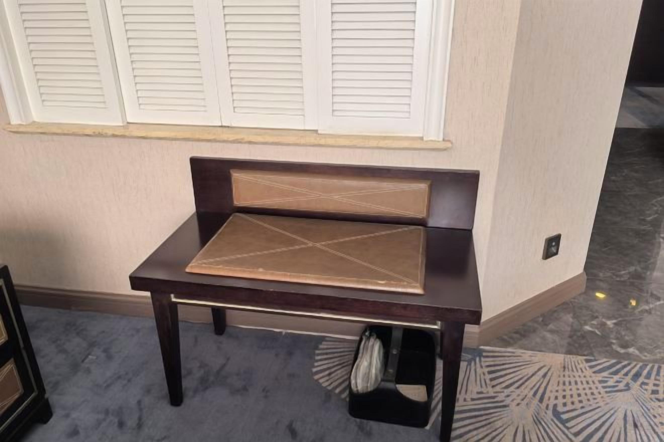 A dark wooden bench with a geometric pattern on the seat, positioned against a wall with a window with white shutters.