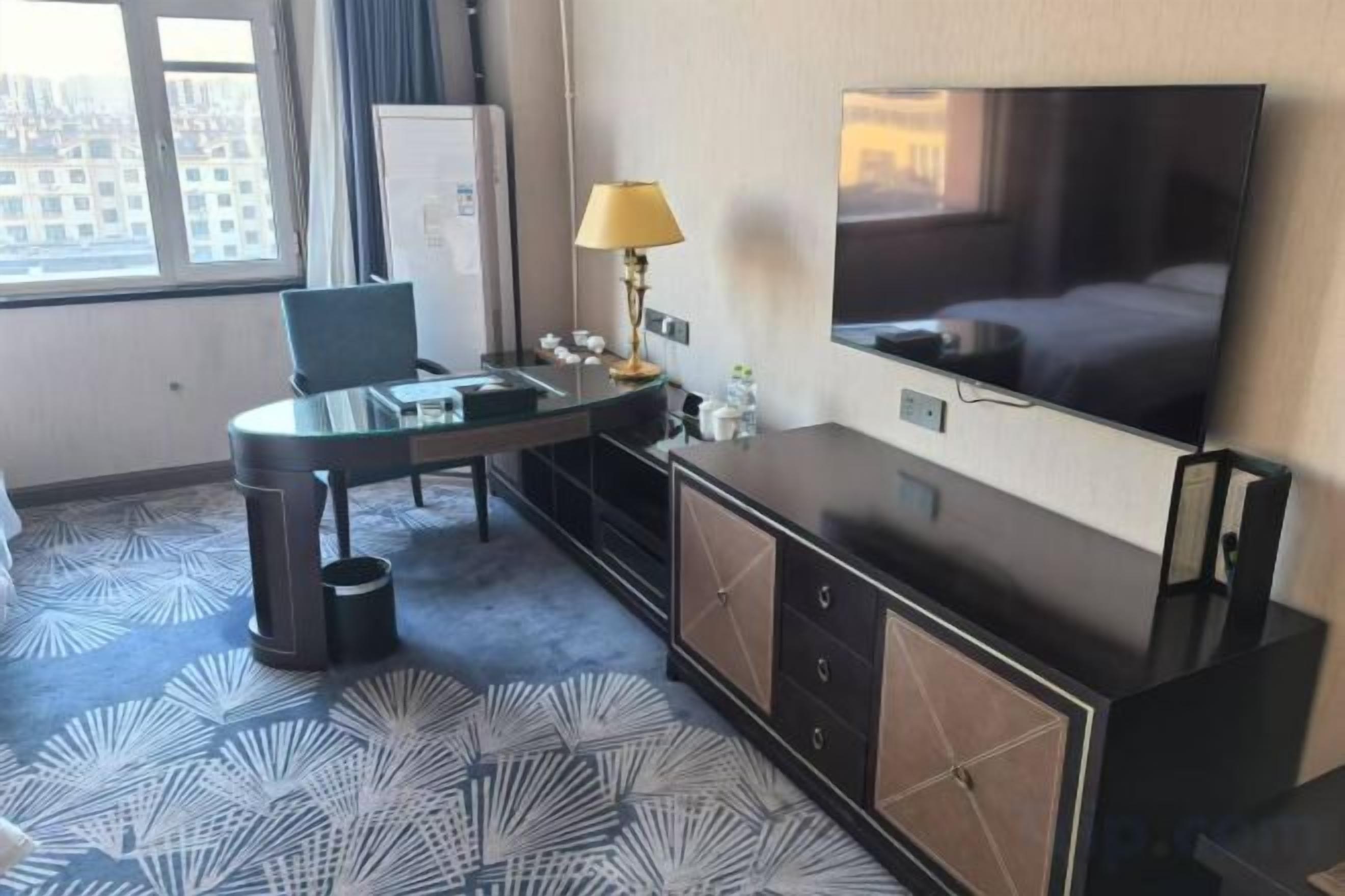 Habitación de hotel con cama, televisor, escritorio con silla, lámpara y vista a la ciudad por la ventana.