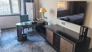 Habitación de hotel con cama, televisor, escritorio con silla, lámpara y vista a la ciudad por la ventana.