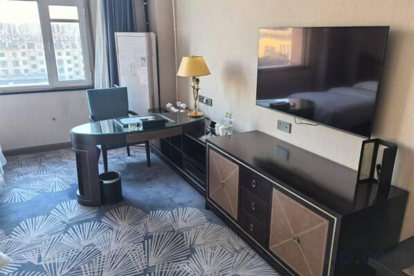 Habitación de hotel con cama, televisor, escritorio con silla, lámpara y vista a la ciudad por la ventana.