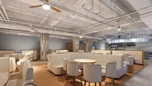 Un espacio lounge moderno con disposición de asientos, una mesa central y una cocina al fondo.