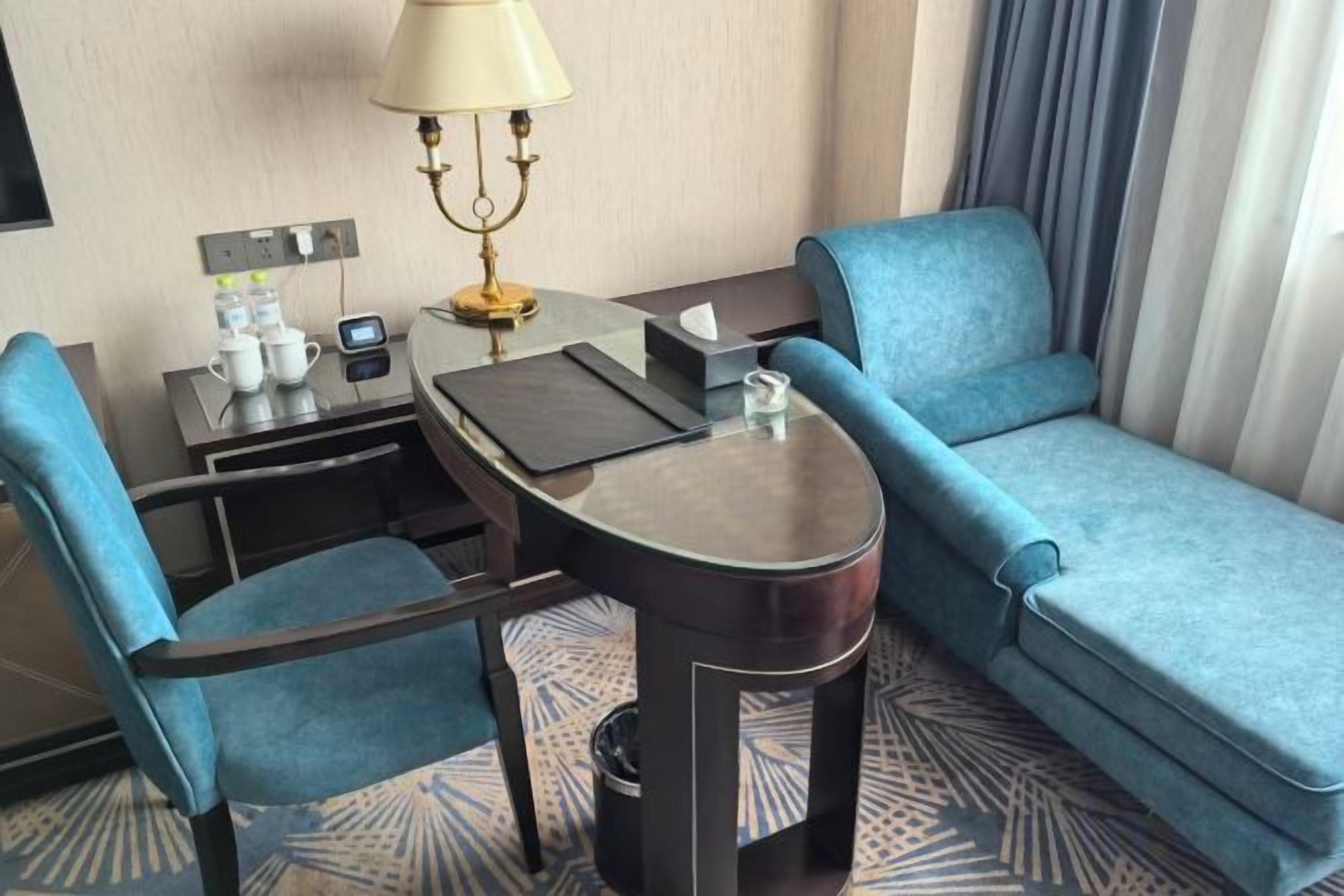 A hotel room with a sofa, a small table with a lamp, a chair, and a desk with a computer and a phone.