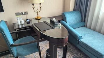 A hotel room with a sofa, a small table with a lamp, a chair, and a desk with a computer and a phone.