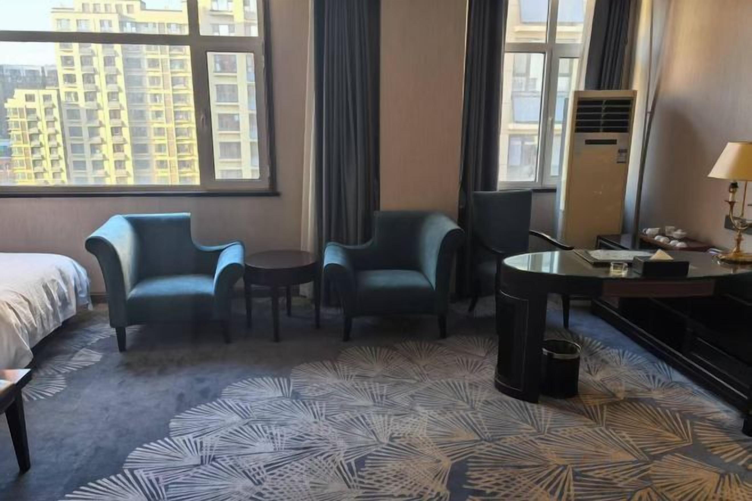 A hotel room with a bed, two armchairs, a small table, a desk with a lamp, and a large window with a city view.